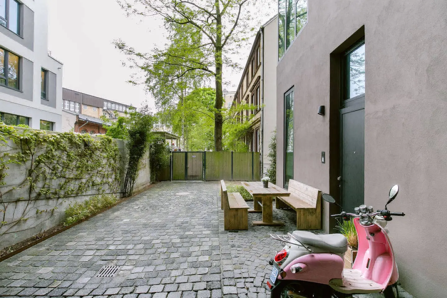 Gepflasterter Hof mit Holztisch, Bänken und rosa Motorroller vor der Loftfassade