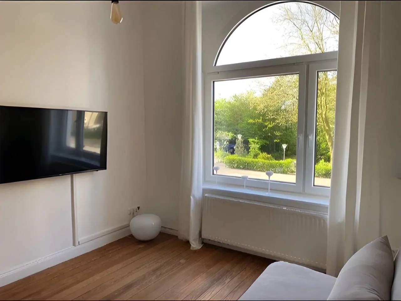 Wohnzimmer mit Bogenfenster, Wand-TV, grauem Schlafsofa und Dielenboden