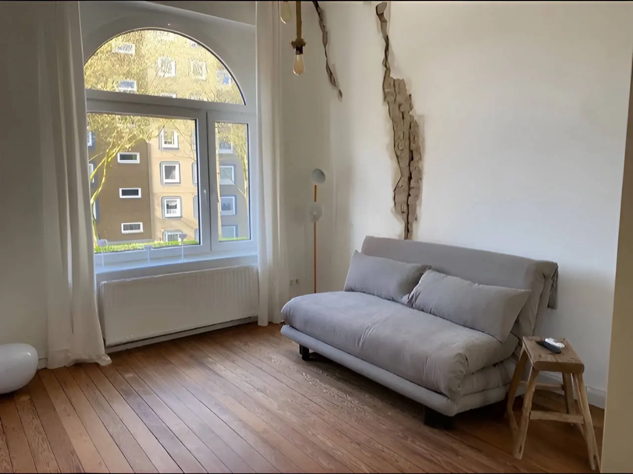 Graues Schlafsofa mit Kissen vor Bogenfenster, Holzhocker und freigelegter Wandstelle