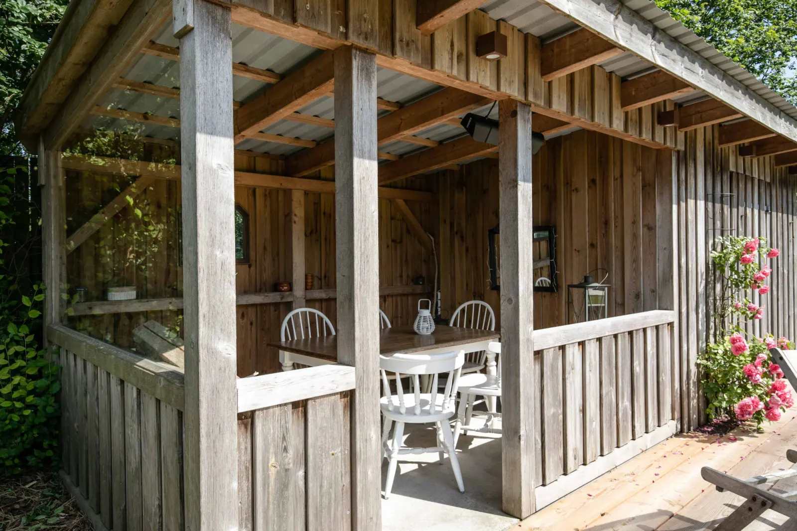 Überdachter Holzpavillon mit weißen Stühlen und Tisch, rosa Rosen am Pfosten