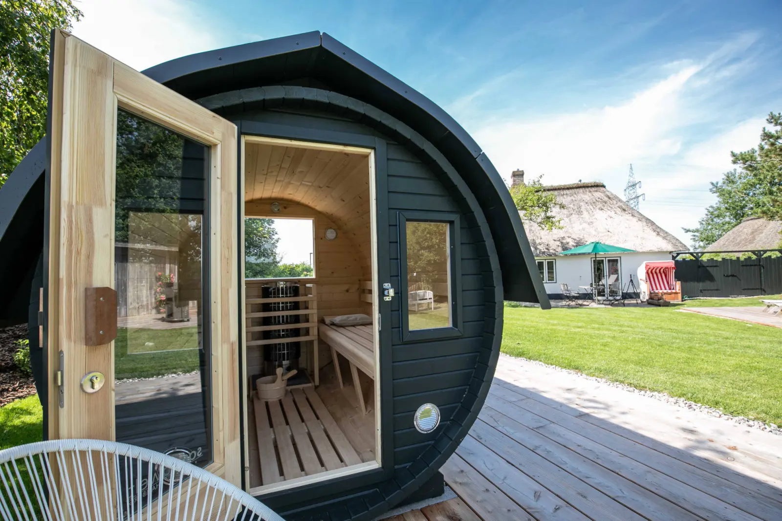Fasssauna mit geöffneter Glastür, Blick in die Saunakabine, dahinter das Haus