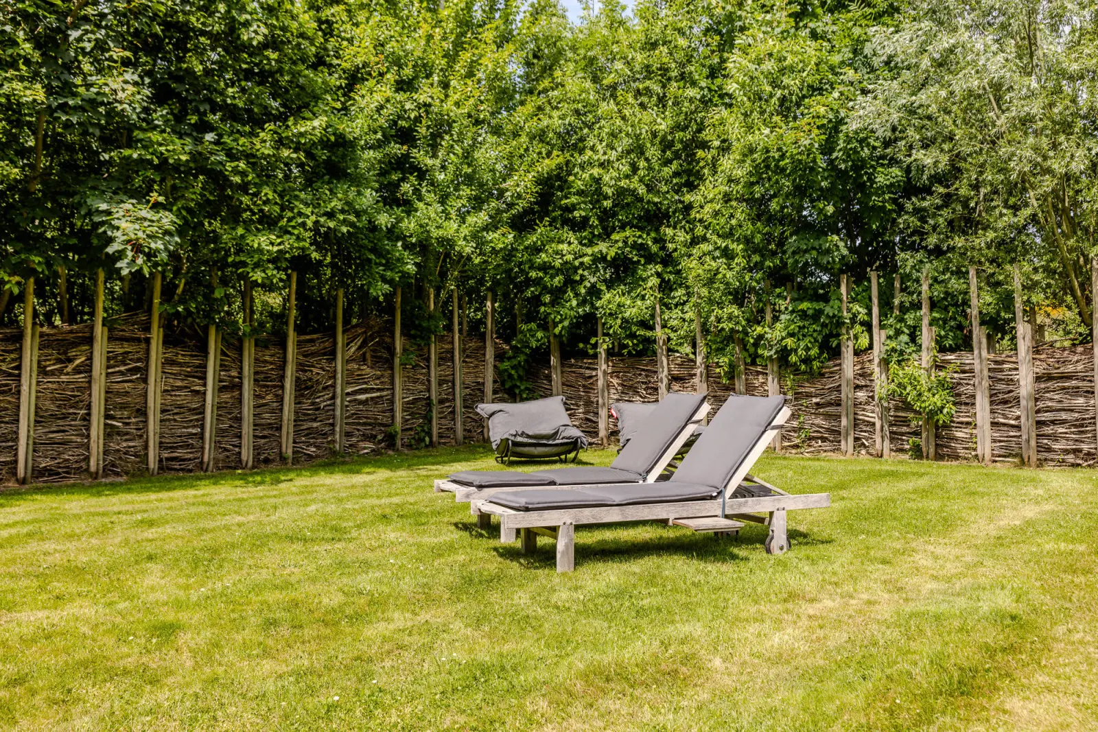 Zwei Sonnenliegen auf der Rasenfläche vor einem Weidenzaun im Garten