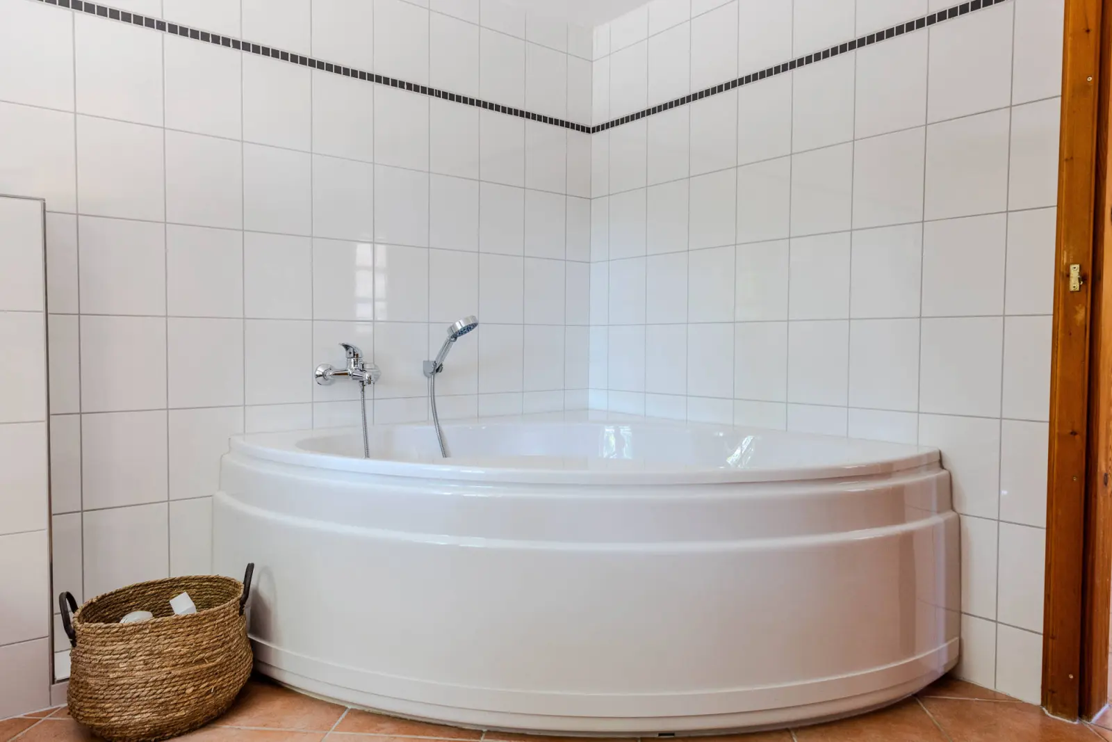 Weiße Eckbadewanne mit Armatur und Korb im gefliesten Bad
