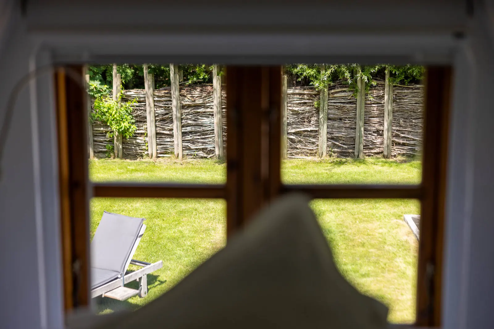 Blick aus dem Fenster auf Weidenzaun und Sonnenliege im Garten