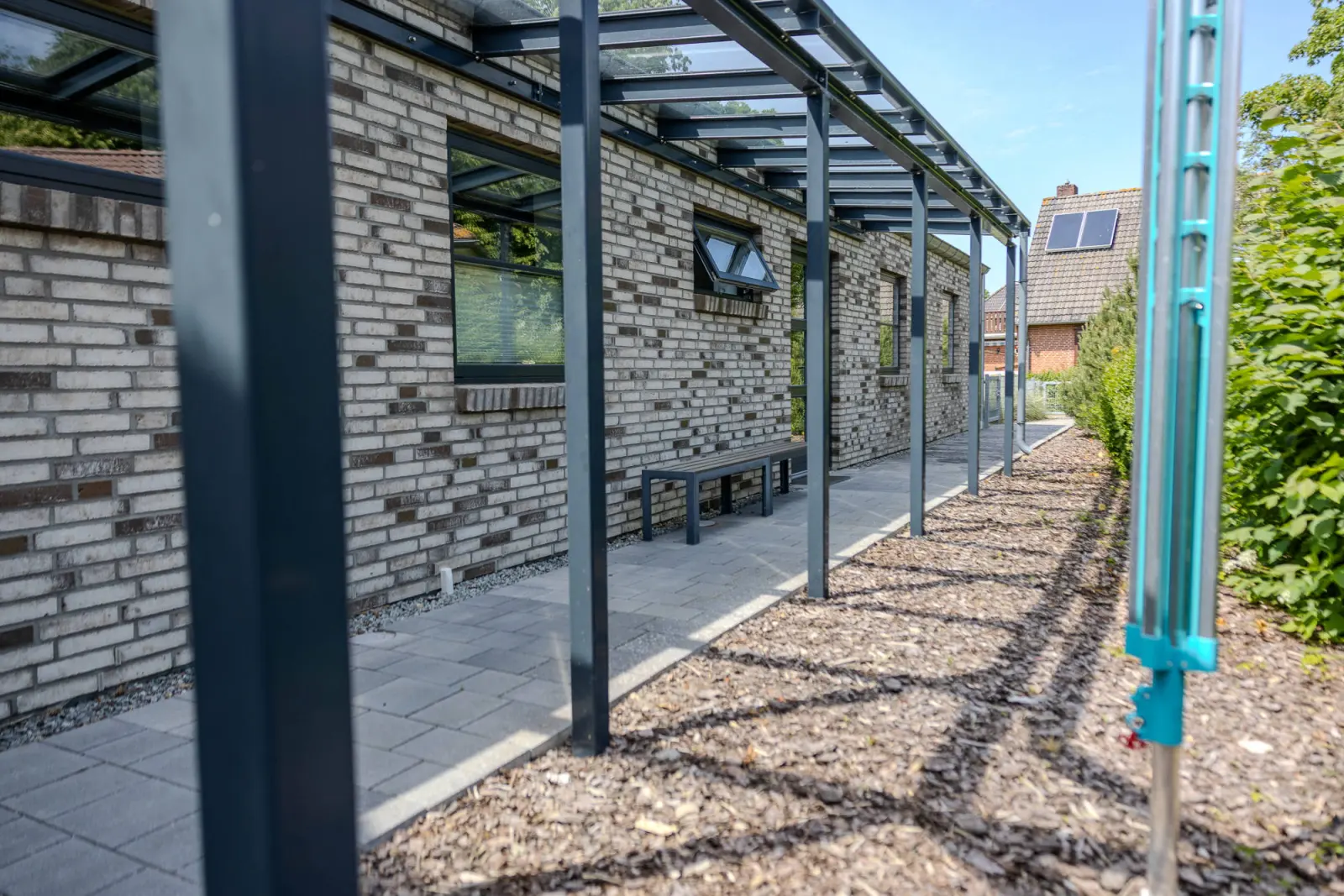 Glasüberdachter Weg entlang der Hauswand mit Holzhackschnitzeln und Kiefer