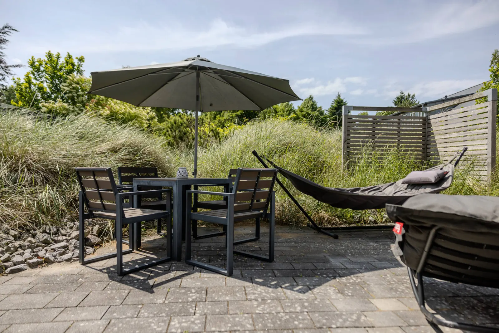 Gartentisch mit Stühlen und Hängematte vor Dünengras und Holzzaun