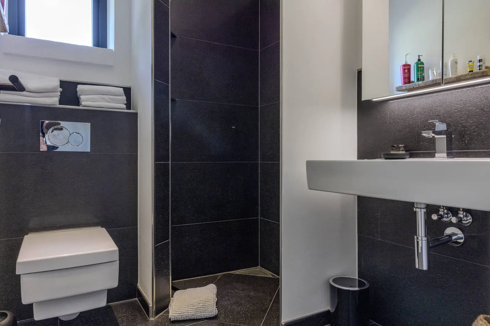 Bad mit Wand-WC, Waschbecken und dunkel gefliester Dusche