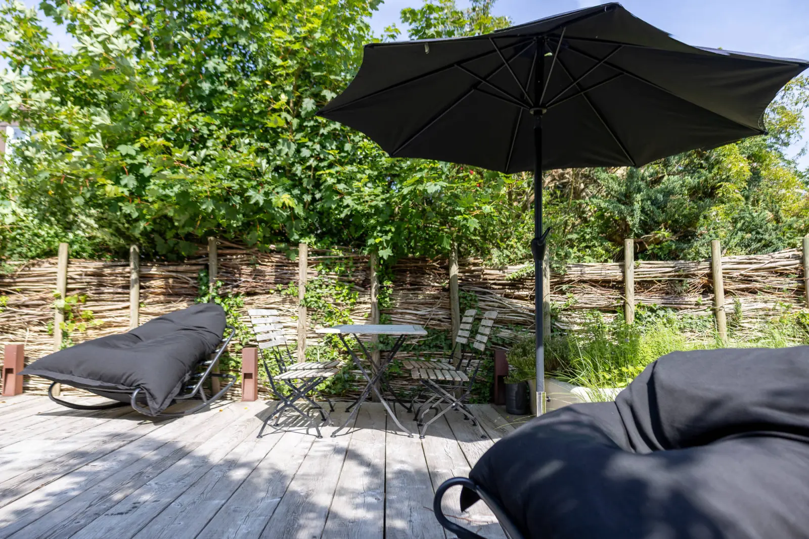 Holzdeck mit schwarzem Sonnenschirm, Gartenstühlen und Sitzsack vor Weidenzaun