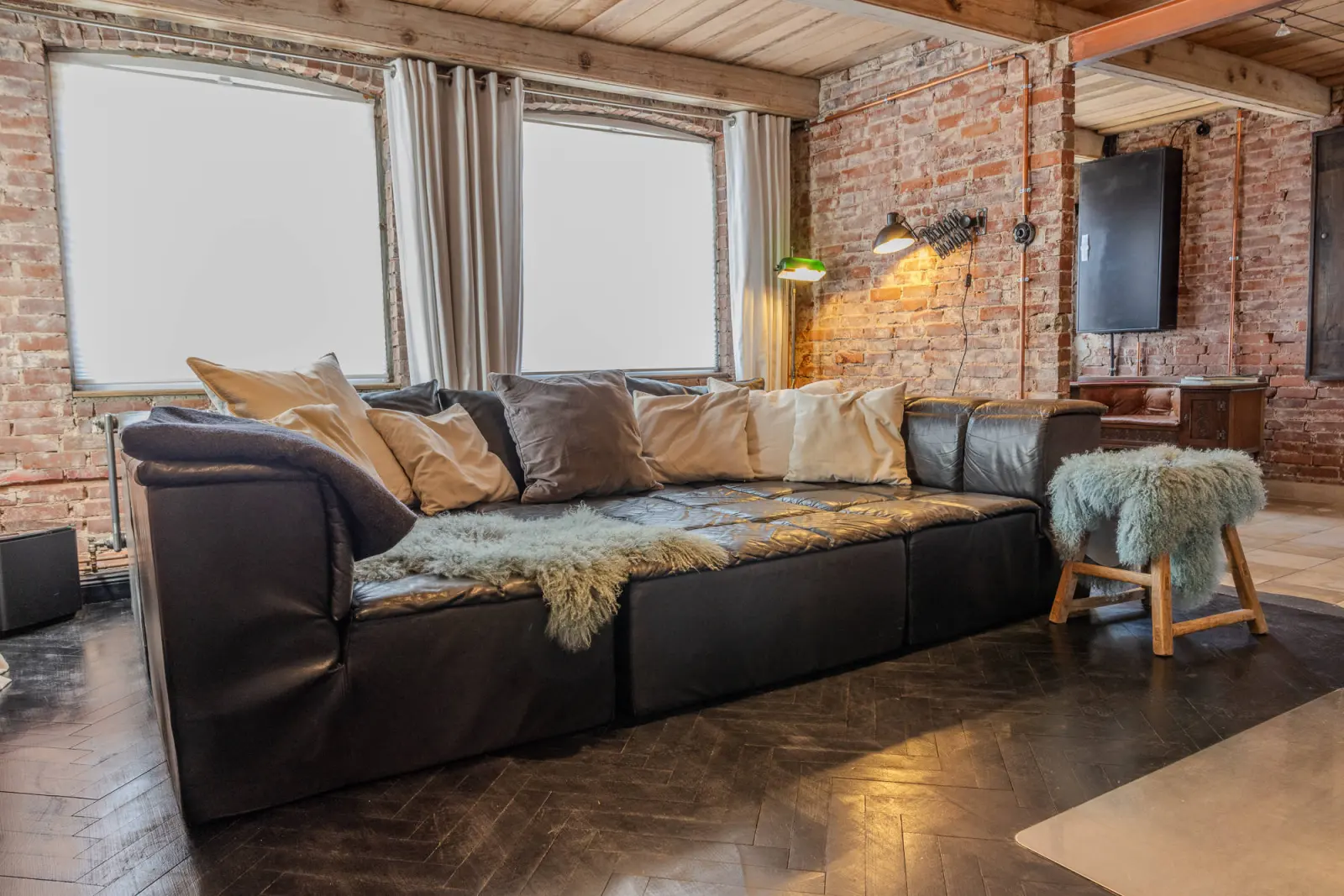Wohnzimmer mit schwarzem Ledersofa, Fellhocker und Wand-TV vor Backsteinwand