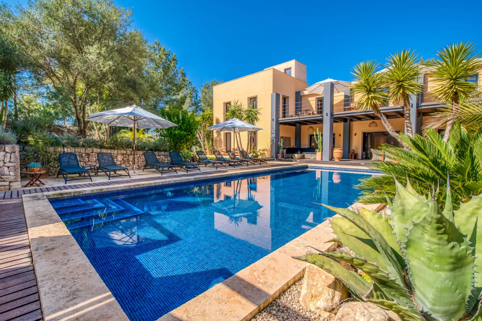 Pool mit Mosaikfliesen, Liegen und Agave, dahinter die Finca im Sonnenlicht