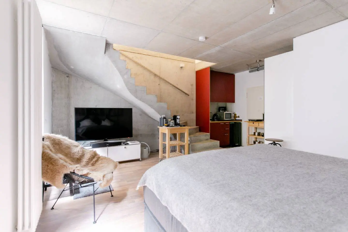 Bett im Loft mit Fellsessel, Betontreppe und kleiner Küchenzeile mit roter Wand