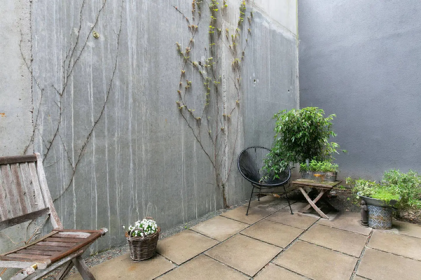 Terrasse mit Steinplatten, Holzstuhl, Korb mit Blumen und Rankpflanzen an der Betonwand