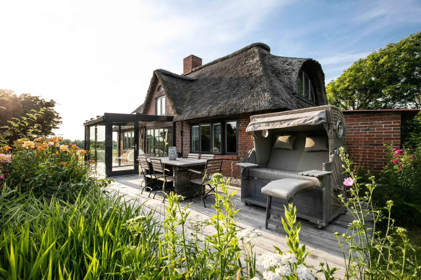 Holzterrasse mit Esstisch, Strandkorb und Wintergarten, davor Taglilien im Beet
