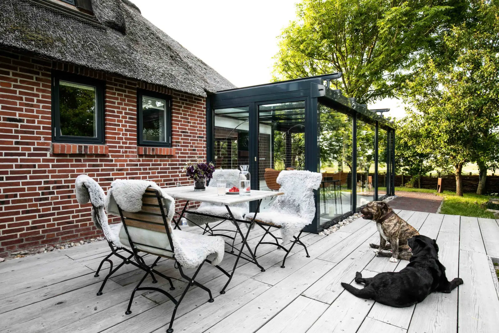 Terrasse mit Klappstühlen, Schaffellen, Esstisch und zwei Hunden vor dem Wintergarten