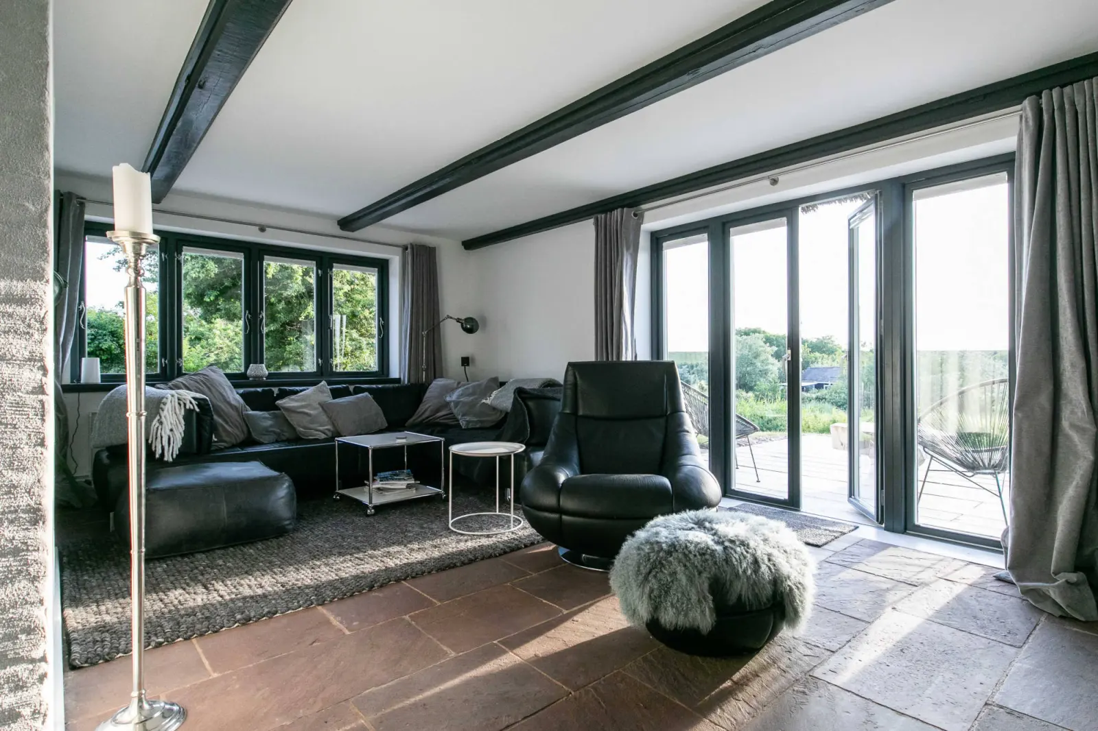 Wohnzimmer mit schwarzem Ledersofa, Sessel, Holzbalken und Terrassentüren