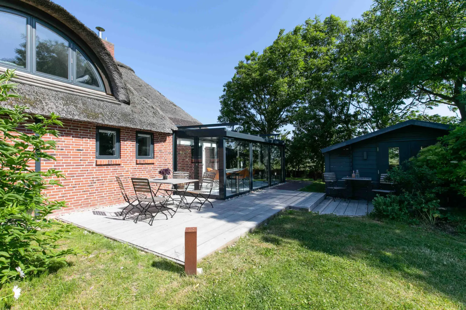 Terrasse mit Gartenmöbeln und Wintergarten, daneben ein dunkles Nebengebäude