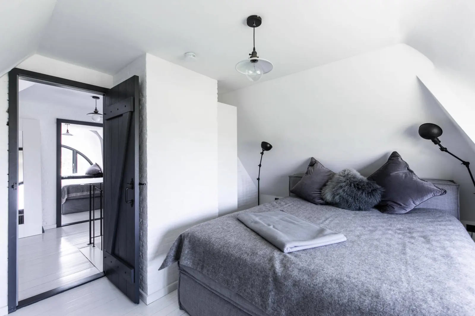 Doppelbett mit grauen Kissen, schwarzer Holztür und Stehlampen an der Wand