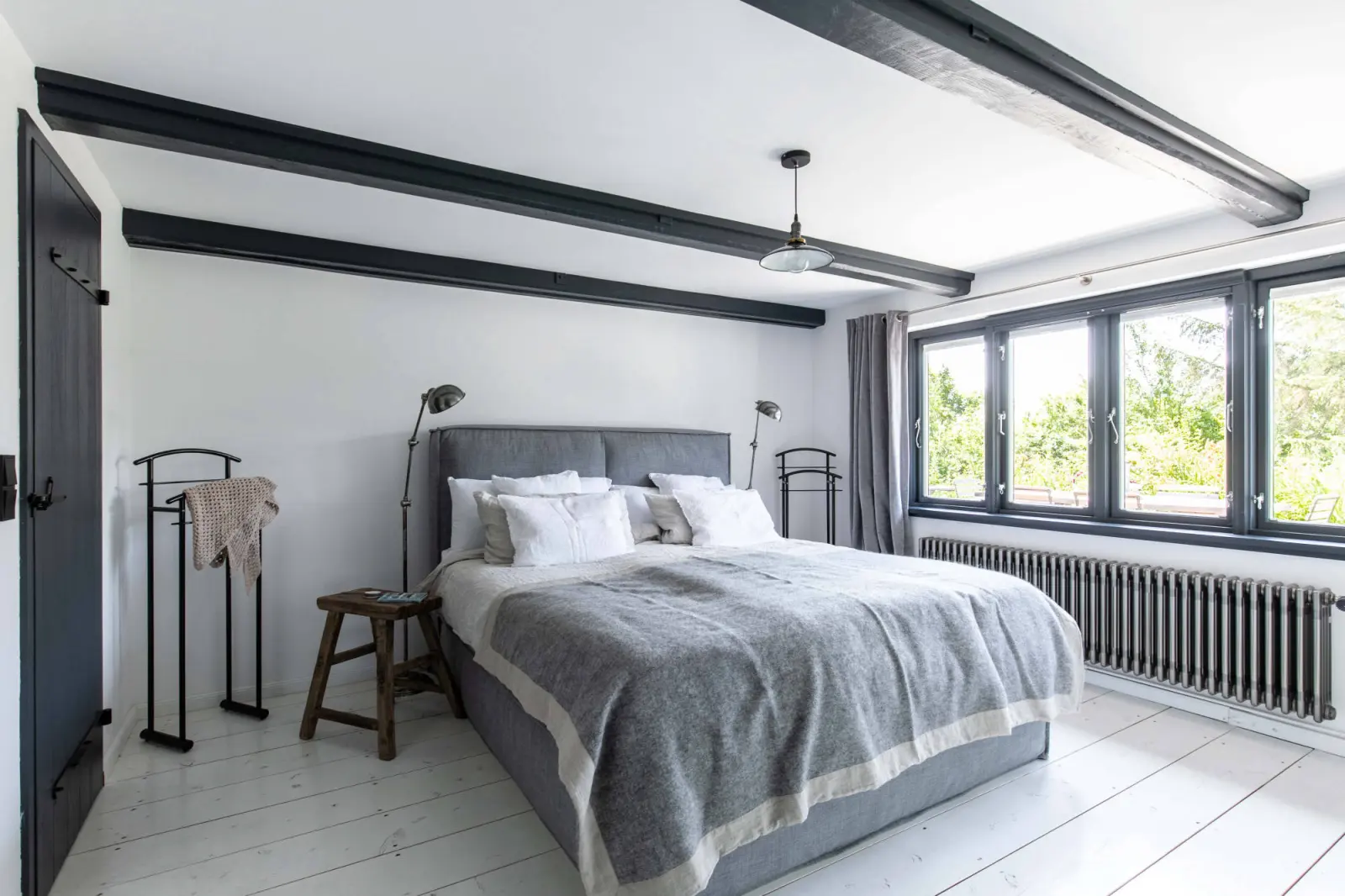 Schlafzimmer mit grauem Polsterbett, Holzbalken und Fenstern zum Garten