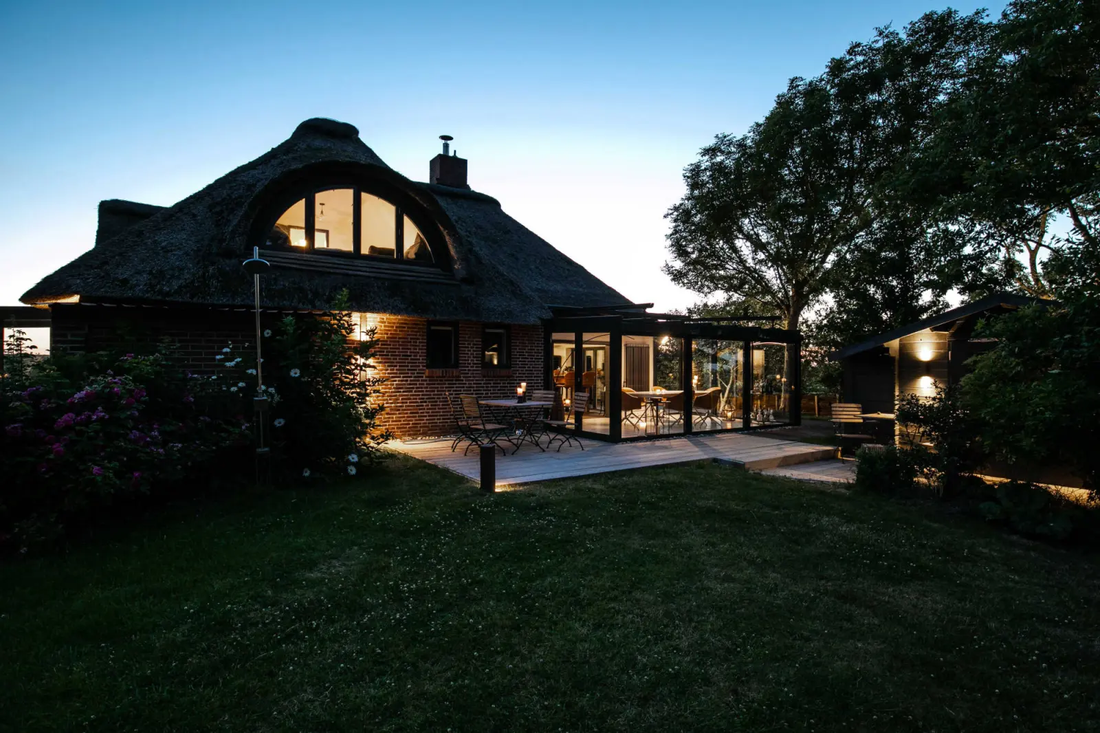 Beleuchtetes Reetdachhaus in der Dämmerung mit Rundgaube und Wintergarten